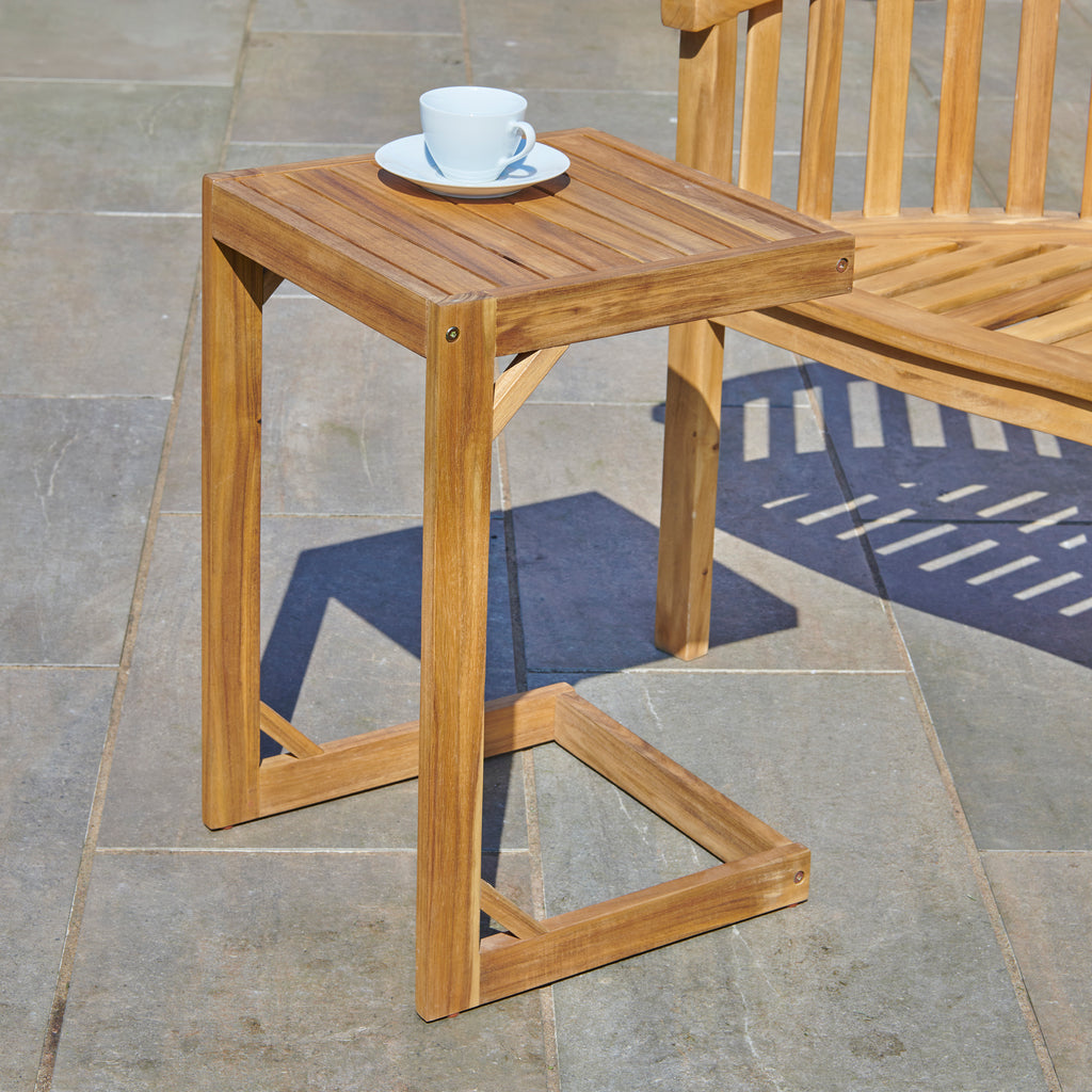 An Acacia Outdoor Side Table, made from FSC Acacia hardwood, holds a cup and saucer and is placed beside a wooden bench on a stone patio.